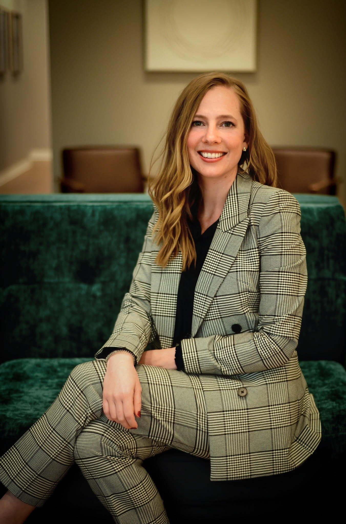 Smiling woman with long blonde hair seated on a green velvet sofa, wearing a checkered suit and black top.